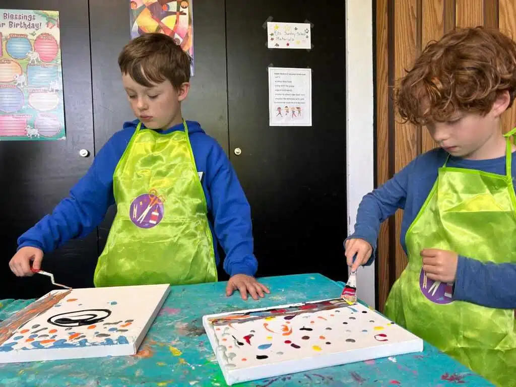Two children painting at a table, wearing aprons.