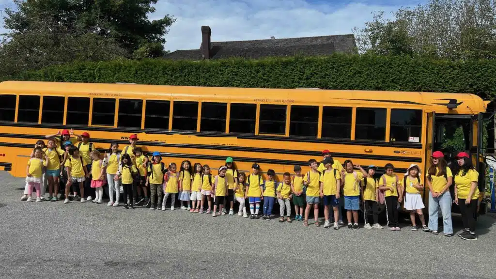 A large group of funderland camp children in yellow shirts stands in front of a yellow school bus with their teachers