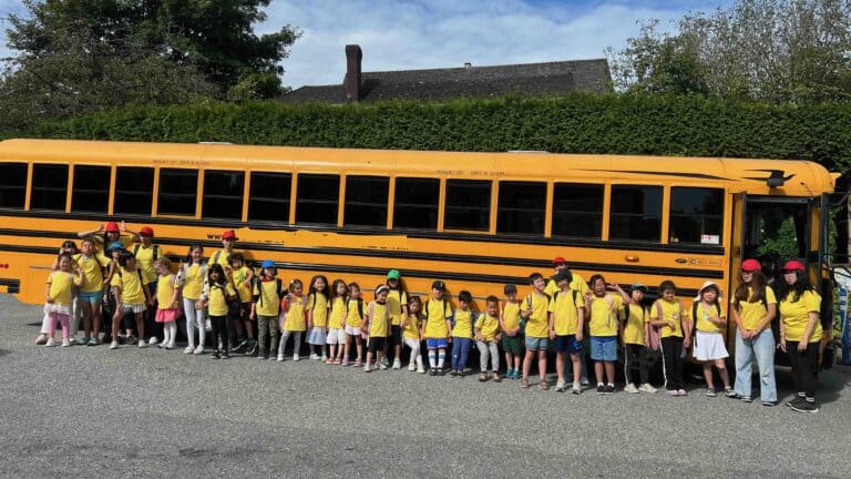 A large group of funderland camp children in yellow shirts stands in front of a yellow school bus with their teachers