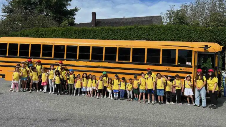 A large group of funderland camp children in yellow shirts stands in front of a yellow school bus with their teachers